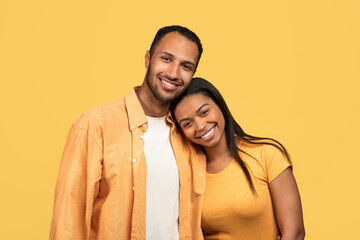 Loving young African American couple cuddling, looking at camera and smiling on yellow studio background