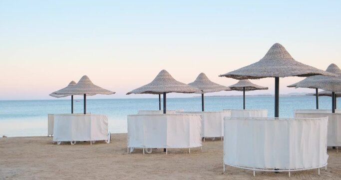 Summer morning on the coast sea ocean empty beach with chairs straw umbrellas and wind windbreaker. Clean water, sky and sun on the background. Sand coastline, landscape panorama vacation holiday time