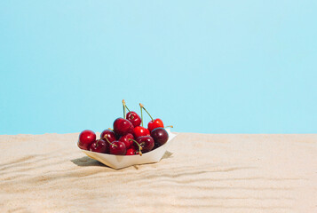 Paper boat filled with ripe cherries on the beach sand.  Minimal summer vacation concept.