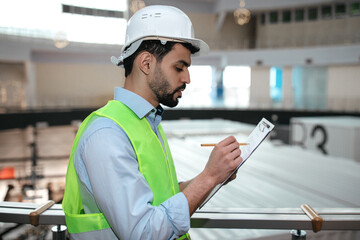 Thoughtful millennial islamic man engineer in protective uniform and hard hat with beard makes notes in tablet