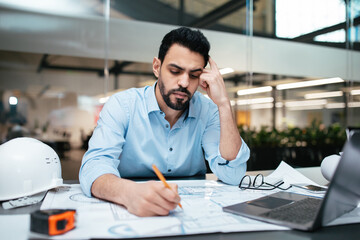 Sad serious pensive attractive millennial arab man engineer with beard works with project drawings