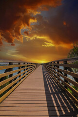 Obraz na płótnie Canvas a long brown wooden bridge over a lush green marsh surrounded by vast blue ocean water with powerful clouds at sunset at Bolsa Chica Ecological Reserve in Huntington Beach California USA