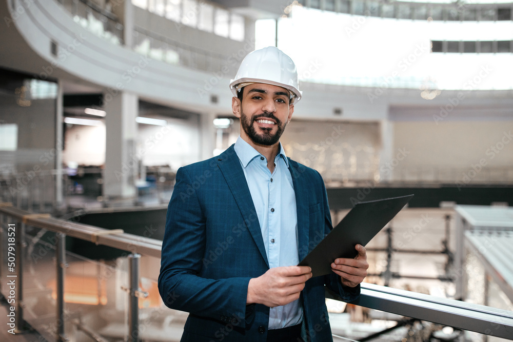 Wall mural smiling confident handsome millennial arab male engineer businessman in protective helmet, suit with