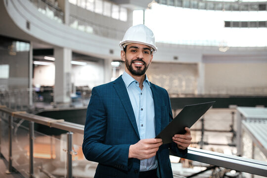 Smiling confident handsome millennial arab male engineer businessman in protective helmet, suit with beard with tablet