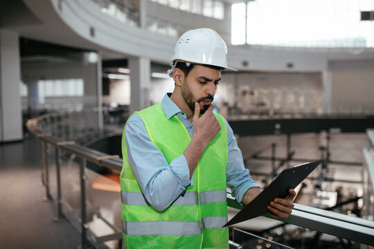 Pensive young islamic engineer builder touches his beard in protective clothing and hard hat looks at tablet
