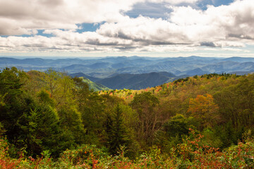 Layers of light and shade and layer after layer of Mountain ridges fall colors of red, yellow orange, and evergreens, clouds from white to dark, with some sun light Horizontal Photo    