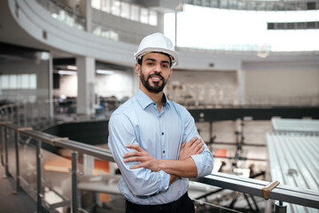Cheerful confident millennial middle eastern male engineer in protective helmet with beard crossed...