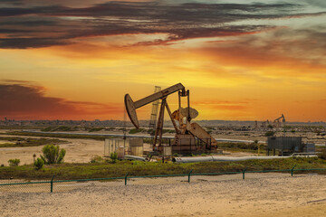 a rusty metal oil rig surrounded by dirt and green plants and grass with powerful clouds at sunset...