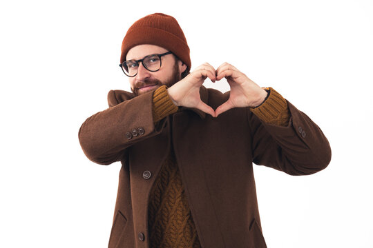 Handsome caucasian hipster man in elegant brown autumn outwear looking at camera and showing heart gesture with both his palms over white background. High quality photo