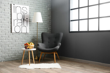 Fruit basket on table, armchair, floor lamp and painting with word INSPIRATION on grey brick wall in living room