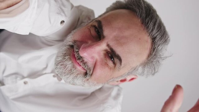 Vertical studio closeup shot of a content happy caucasian bearded man with gray-hair waving at camera and smiling. High quality 4k footage