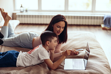 Small Caucasian cute boy with beautiful mother lying together on bed with laptop. © Kostiantyn
