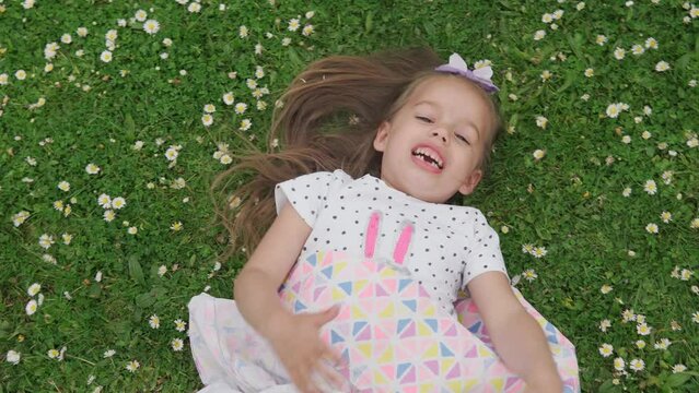 Little happy child girl in dress with bow on head laying on green lawn in the park. Summer time, nature, Dreams, Lifestyle Concepts. Smilling Baby Face close up. emotional child grimaces at camera