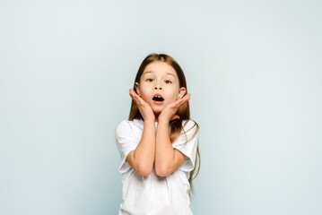 A little girl holds her hands near her face and is surprised on a blue background. Children's discounts and promotions.