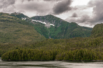 Pacific Coastline, Alaska, USA - July 16, 2011: Canadian Rocky mountain under heavy cloudscape. Detail shows muddy waterfall and snow patches on heavy forested green flanks.