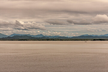 Pacific Coastline, Alaska, USA - July 16, 2011: Canadian Rocky mountains under heavy cloudscape with green forested coastline. Small fishing vessel om brown Pacific water.
