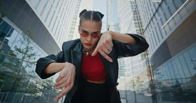 Female model wearing black jacket and cool red eye glasses dancing seductively with hands near high rise skyscrapers in downtown district. Creative urban style woman modern city background