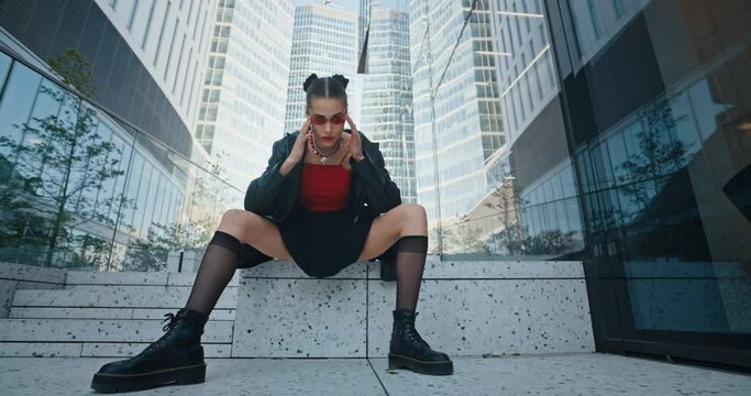 Sexy young female wearing black jacket short skirt stockings and red eye glasses seductively posing sitting on stairs near glass skyscrapers in downtown district. Cheeky urban style fashion model