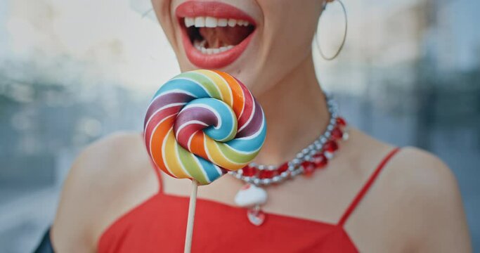 Close-up gorgeous happy woman seductively licks big multi-colored lollipop and smiles, stylish teen model wearing cool red eye glasses posing with colorful candy standing in modern city street