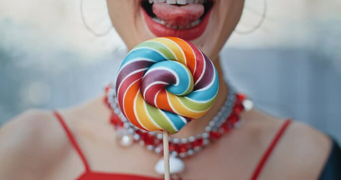 Close-up attractive happy woman seductively licks big multi-colored lollipop and smiles, stylish teen model wearing cool red eye glasses posing with colorful candy outdoors in city street