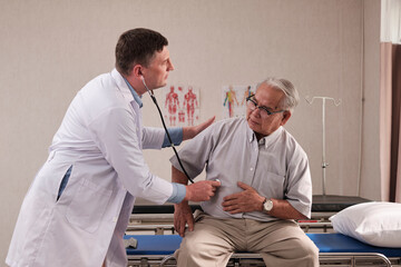 Obraz premium Caucasian male doctor in uniform health checks illness senior patient with stethoscope, painful stomach ache in bed of emergency room at hospital ward, elderly medical clinic examination consultant.