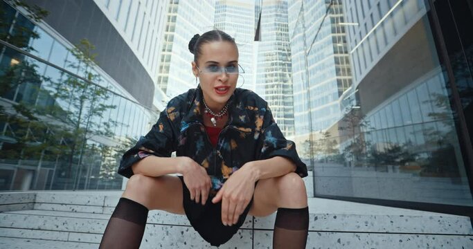 Urban style cheeky female wearing short colored windbreaker jacket and futuristic glasses rapping expressive while sitting on stairs in modern downtown overlooking high-rise glass skyscrapers