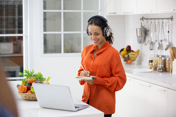 Smiling young aspiring female chef looking and writing recipe of new dish on laptop while listening...