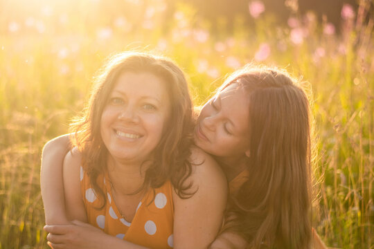 Portrait of a smiling happy Caucasian mother with her daughter in orange dresses on the background of a sunset field. An adult daughter kisses her mother. The concept of the Mother's Day holiday.