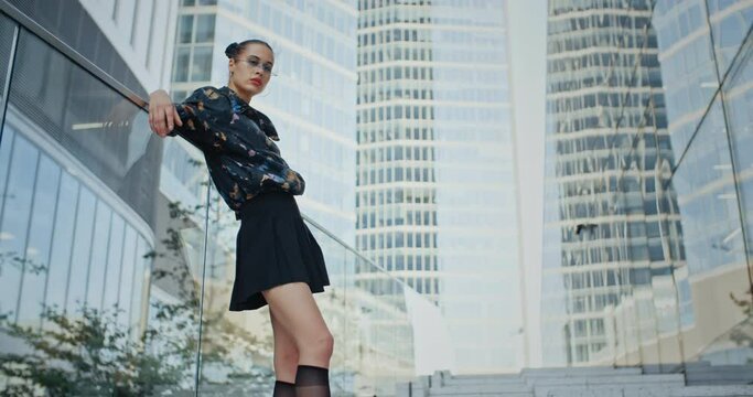 Urban style seductive girl wearing colored jacket short skirt and futuristic glasses posing leaning back against glass parapet overlooking high rise glass skyscrapers. Beautiful female fashion model