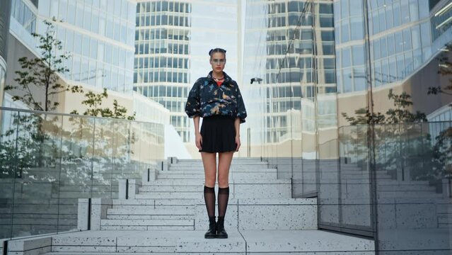 Urban style slim girl wearing short skirt futuristic glasses boots stockings standing still and looking at camera overlooking modern cityscape high rise skyscrapers. Attractive female fashion model