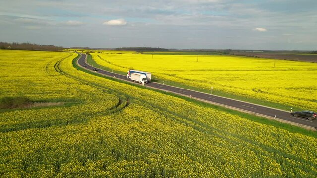 AERIAL: Cargo trucks and cars driving on motorway road passing by yellow fields. Running traffic on beautiful countryside asphalt roadway surrounded with flowering brassica rapa agricultural fields.