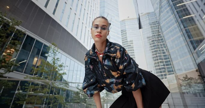 Wide angle urban style seductive sexy woman wearing jacket short skirt futuristic glasses posing looking at camera overlooking high rise skyscrapers modern downtown. Female fashion model