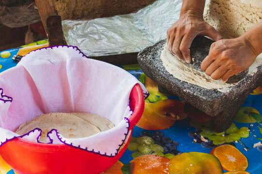 Mujer mexicana torteando maza de maíz en un metate y una estufa de leña para hacer tortillas caseras  