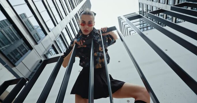 Wide angle urban style sexy young woman wearing colored jacket and cool red glasses standing on stairs leaning on railing and looks into camera seductively overlooking high modern buildings