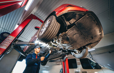 Young car mechanic at repair service station inspecting car wheel and suspension detail of lifted automobile. Bottom view.