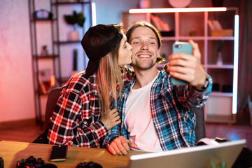 Selfie time. Cheerful guy taking photo on smartphone with his girlfriend in livivng room. Romantic Caucasian couple sitting at table, loving woman kissing her boyfriend.