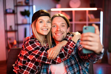 Smiling beautiful family couple posing for selfie on cellphone. Happy young hispanic woman showing funny mobile apps to loving husband, sitting together at table in modern living room, tech addiction.