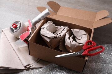 Open box with wrapped items, adhesive tape, scissors, paper and bubble wrap on wooden table