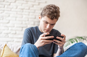 A teenage boy plays games on a smartphone with a focused expression. Dependence on the phone and online games. Close-up, bottom view. A young man with a phone in his hands sits on the couch.