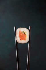 Chopsticks hold a sushi roll with salmon close-up on a dark background. Concept Japaness food