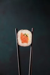 Foto op Plexiglas Chopsticks hold a sushi roll with salmon close-up on a dark background. Concept Japaness food © Kufotos