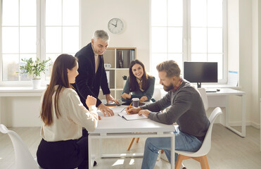 Group of positive people dressed in casual clothes discuss plan for cooperation on common project in modern coworking. People write their ideas in notebook sitting at table in bright office.