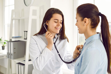 Young friendly nurse listens to woman's heart and breath through stethoscope on her chest. Doctor listens to heart rate of female patient during medical examination at clinic. Medicine concept.