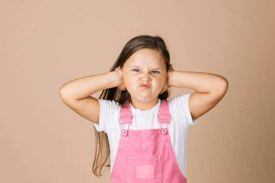Portrait photo of child closing ears with hands not hear what others say with frowny-face with shining eyes looking at camera wearing bright, pink jumpsuit and white t-shirt on beige background.