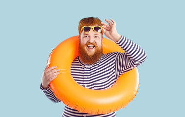 Funny fat man with lifebuoy around his neck, waist and sunglasses isolated on light blue background. Cheerful bearded chubby man with orange inflatable circle lifting goggles funny looking at camera.