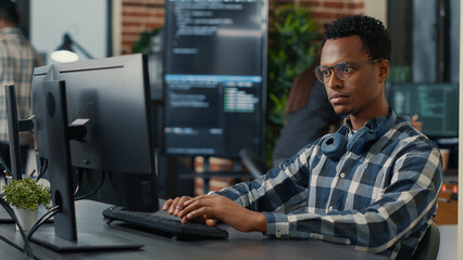 Portrait of focused programer writing code fixing glasses and smiling sitting at desk in it startup. Coder programming user interface with team of programmers working in the background.