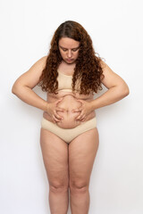 Portrait worried plump woman with curly hair wearing beige underwear, squeezing excess fat of belly, bowing head, looking at stomach on white background. Weight loss, obesity, diet, health problems.
