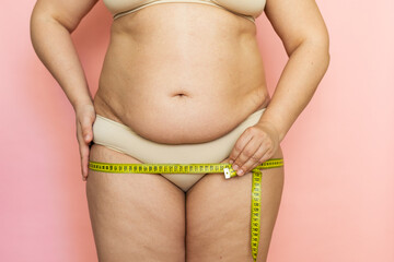 Cropped photo of naked overweight woman body, holding and measuring by roulette tape her hips,...