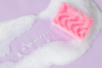 Pink sponge with detergent foam on purple background, close up. Cleaning concept