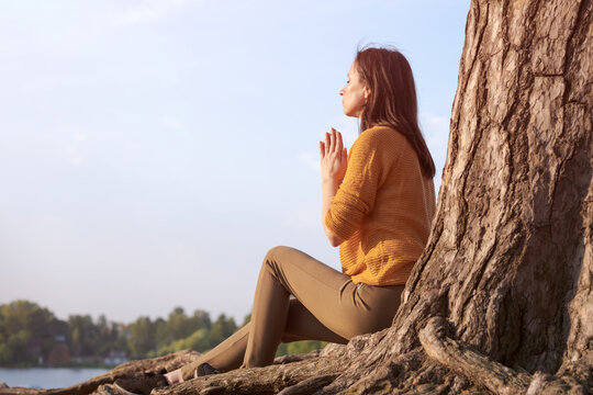 Peaceful woman holds her palms in front her in prayer against backdrop trees on the lake in the rays of the setting sun. Meditating or praying. Caucasian woman believes in a higher power